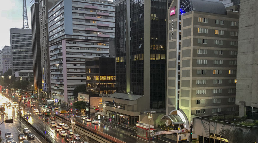 #PraCegoVer Vista aérea da cidade de SãoPaulo com a Avenida Paulista no canto inferior esquerdo da foto e
o hotel IBIS do lado dirito