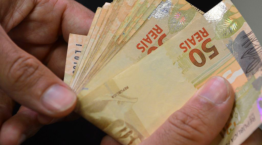 #PraCegoVer Mãos masculinas segurando um maço de notas de cinquenta reais