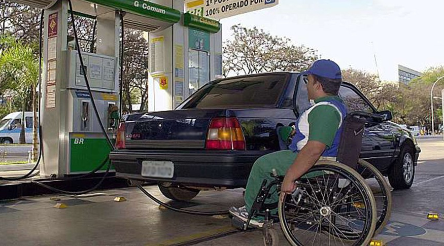 Em primeiro plano, homem preto em cadeira de rodas trabalha como frentista em um posto de gasolina, abastecendo um carro. Ele veste uniforme com calça verde, camisa verde, branca e azul. Ao fundo, vê-se a bomba de gasolina que está sendo utilizada.