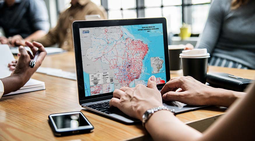 #PraCegoVer A foto mostra os braços de uma mulher, do lado direito da imagem, usando o teclado de um computador em que na tela, aparece o mapa das redes 2021. Ao fundo, aparece uma imagem esfumaçada de escritório com algumas pessoas trabalhando.