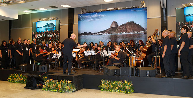 galeria-2 #PraCegoVer apresentação do coral com músicos clássicos em palco preto com imagem do Pão de Açúcar ao fundo