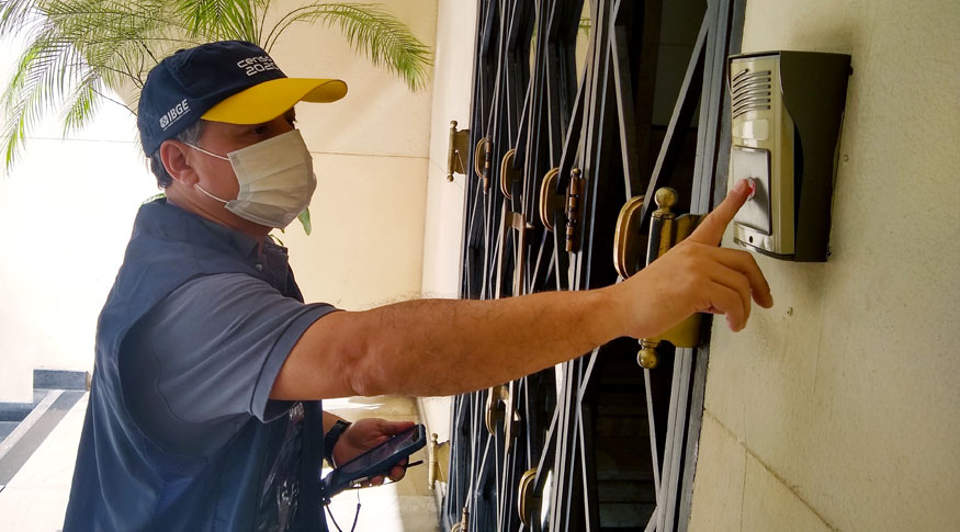 #Pracegover A foto mostra um recenseador do IBGE, com colete e boné de identificaçãotocando a campainha de uma casa.