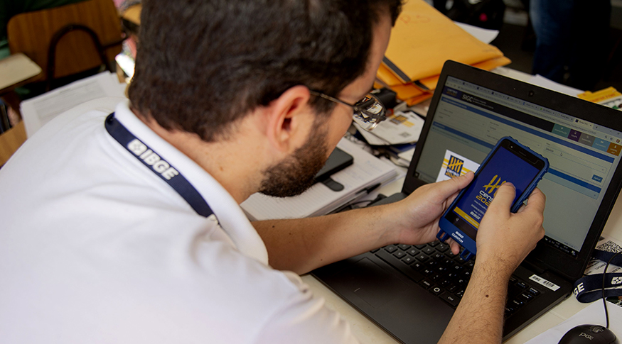 #PraCegoVer A foto mostra um homem sentado, com crachá do IBGE pendurado no pescoço, utilizando um dispositivo móvel de coleta, e na sua frente tem uma mesa e um notebook.